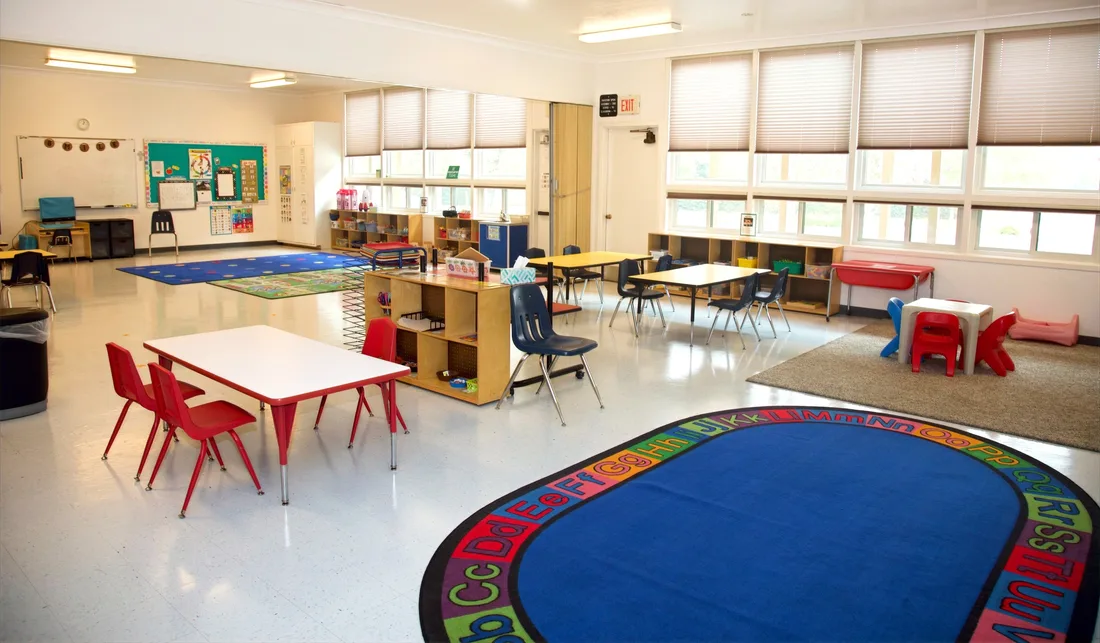 A bright classroom with a large alphabet circle rug, low tables and chairs set for a group, and windows along one wall.
