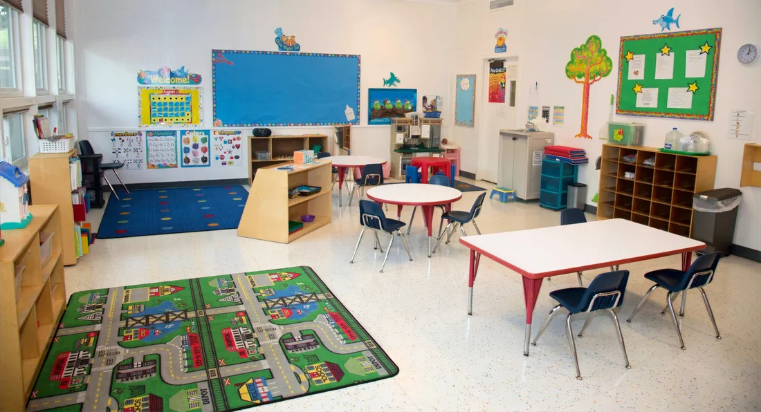 A classroom set for the morning, with a round table, rectangular tables and chairs, cubby shelving and a road rug.