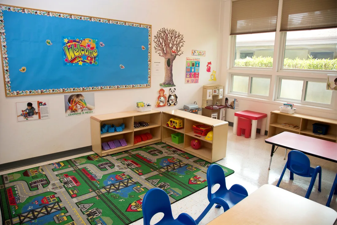 A classroom with a bright Welcome bulletin board, a painted tree on the wall, low shelving of toys and a road rug with small blue chairs.