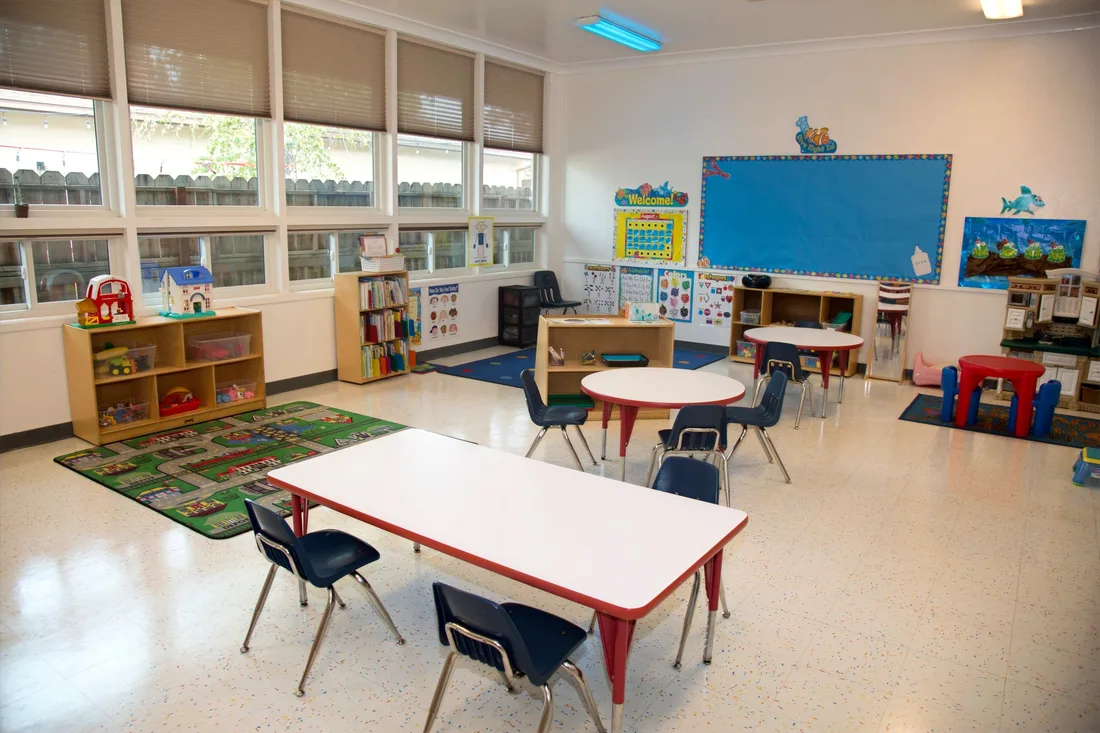 A classroom seen from the doorway, with a wall of windows, a library shelf, a road rug and tables set low.