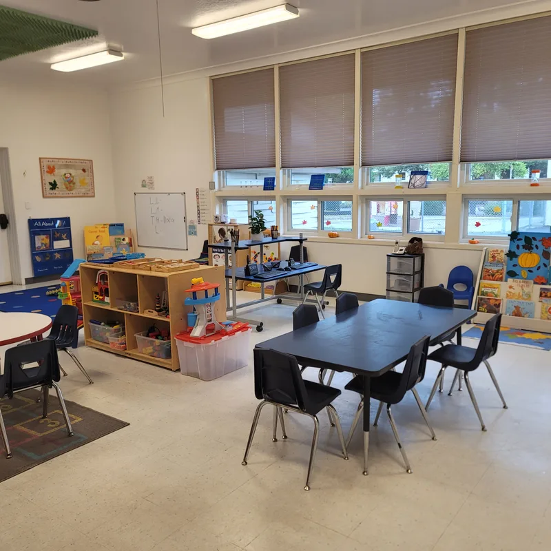 A classroom along its window wall, with building shelves, an easel and tables laid out for a full group.